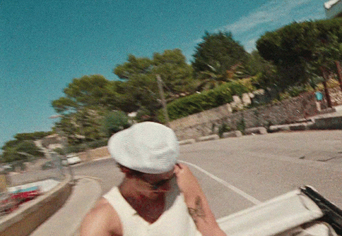 A Man Wearing A White Hat And A White Tank Top Is Walking Down A Street GIF