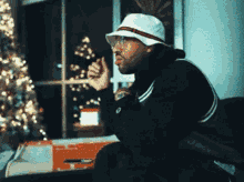 A Man Wearing A White Hat And Glasses Is Sitting In Front Of A Christmas Tree . GIF