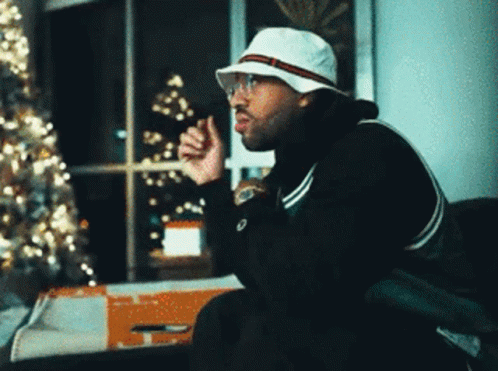 A Man Wearing A White Hat And Glasses Sitting In Front Of A Christmas Tree GIF