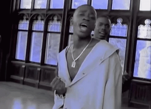 A Man Wearing A White Jacket And A Necklace Sings In Front Of A Window GIF