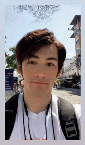 A Man Wearing A White Shirt And A Black Backpack Stands In Front Of A Sign That Says ' T ' On It GIF