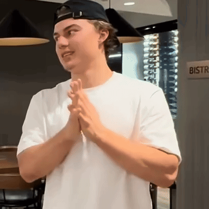 A Man Wearing A White Shirt And A Black Hat Is Standing In Front Of A Bistro Sign . GIF