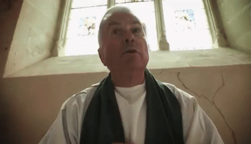 A Man Wearing A White Shirt And A Black Scarf Is Standing In Front Of A Window GIF