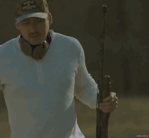 A Man Wearing A White Shirt And A Camo Hat Holds A Gun In His Right Hand GIF