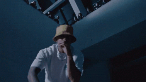 A Man Wearing A White Shirt And A Yellow Hat Is Dancing In A Dark Room GIF