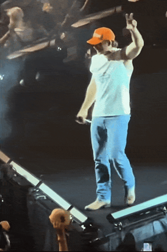 A Man Wearing A White Shirt And An Orange Hat Stands On A Stage Giving A Peace Sign GIF
