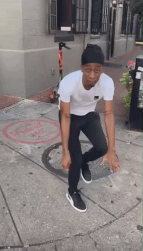 A Man Wearing A White Shirt And Black Pants Is Standing On A Sidewalk GIF