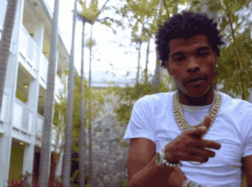 A Man Wearing A White Shirt And Gold Chains Is Standing In Front Of A Building GIF