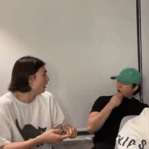 A Man Wearing A White Shirt With A Black Cat On It Is Talking To Another Man Wearing A Green Hat GIF