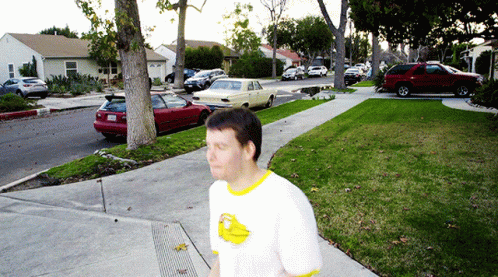 A Man Wearing A White Shirt With A Yellow Duck On It Walks Down The Sidewalk GIF