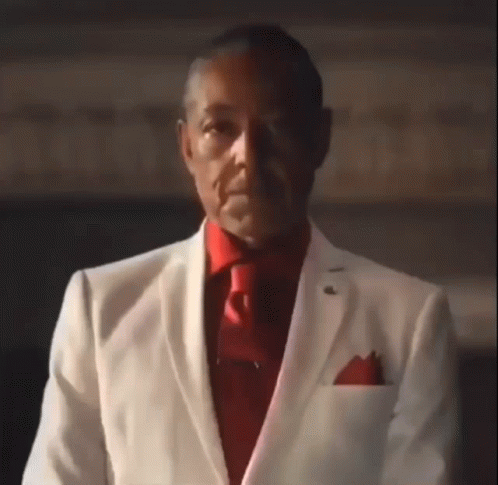A Man Wearing A White Suit And Red Tie Is Looking At The Camera . GIF