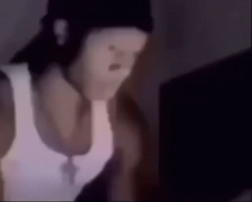 A Man Wearing A White Tank Top And A Black Hat Is Looking At A Laptop . GIF