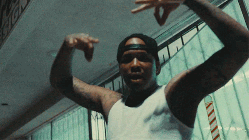 A Man Wearing A White Tank Top And A Black Hat Is Pointing His Fingers GIF