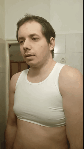 A Man Wearing A White Tank Top Stands In Front Of A White Tiled Wall GIF