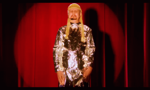 A Man Wearing A Wig And A Silver Jacket Stands On A Stage In Front Of A Red Curtain GIF