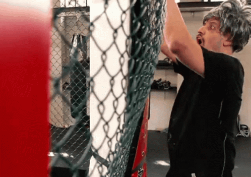 A Man Wearing A Wig Is Standing In Front Of A Chain Link Fence In A Gym GIF
