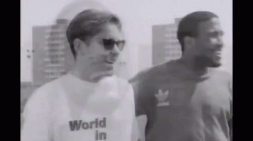A Man Wearing A World In T-Shirt Is Standing Next To Another Man Wearing Sunglasses . GIF