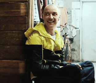 A Man Wearing A Yellow And Black Jacket Is Smiling While Leaning Against A Wooden Wall GIF