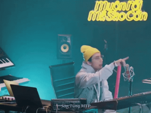 A Man Wearing A Yellow Beanie And Glasses Is Standing In Front Of A Microphone . GIF