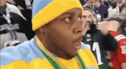 A Man Wearing A Yellow Hat And A Blue And Yellow Sweater With A Green Necklace Around His Neck GIF