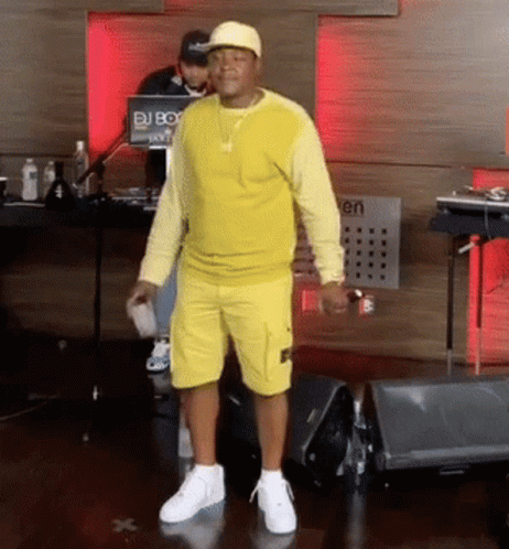 A Man Wearing A Yellow Shirt And Shorts Stands In Front Of A Dj Box GIF