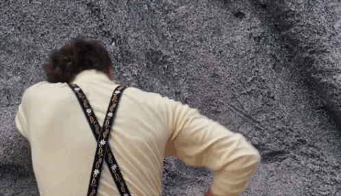 A Man Wearing A Yellow Shirt And Suspenders Is Standing In Front Of A Rock GIF