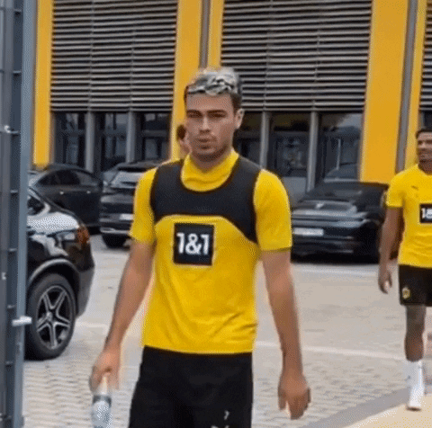 A Man Wearing A Yellow Shirt With The Number 187 On It Is Walking Down A Street Holding A Bottle Of Water . GIF