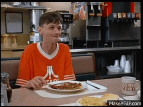 A Man Wearing An Atari Shirt Is Sitting At A Table With A Plate Of Food On It GIF