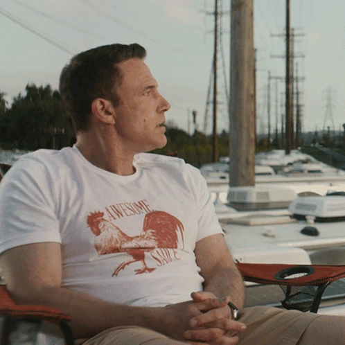 A Man Wearing An Awesome Sauce Shirt Sits On A Chair GIF