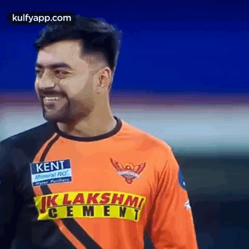 A Man Wearing An Orange And Black Jersey With A Yellow Stripe On The Front Is Smiling . GIF