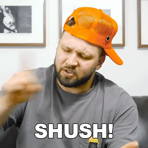 A Man Wearing An Orange Hat And A Grey Shirt Says " Shush " GIF