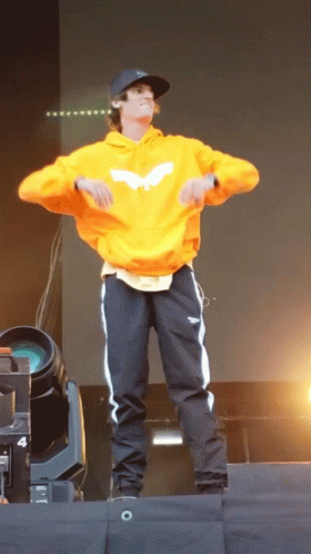 A Man Wearing An Orange Hoodie And Black Pants Is Dancing On A Stage GIF