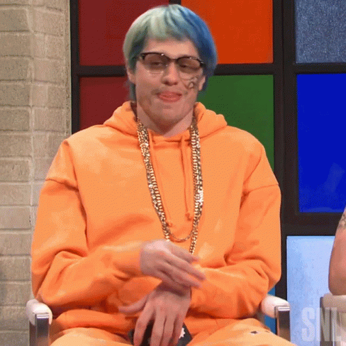 A Man Wearing An Orange Hoodie And Sunglasses Is Sitting In Front Of A Snl Sign GIF