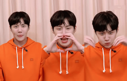 A Man Wearing An Orange Hoodie Is Making A Heart With His Hands GIF