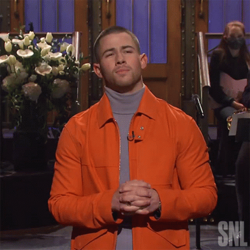 A Man Wearing An Orange Jacket And A Grey Turtleneck Is Standing In Front Of A Snl Sign GIF