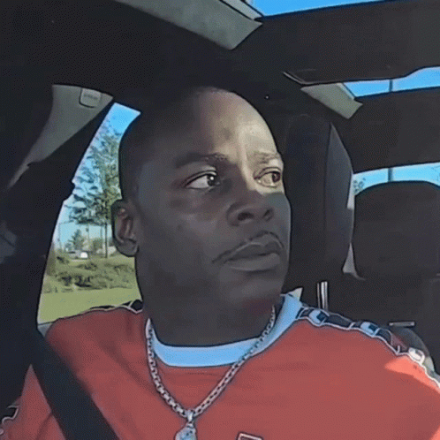 A Man Wearing An Orange Shirt And A Necklace Is Sitting In A Car With His Eyes Closed GIF