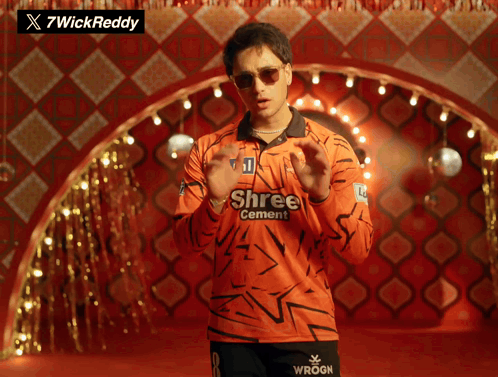 A Man Wearing An Orange Shree Cement Shirt Stands In Front Of A Checkered Wall GIF