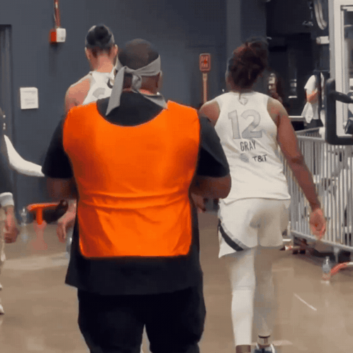A Man Wearing An Orange Vest Walks Behind A Woman Wearing A White Jersey With The Number 12 On It GIF
