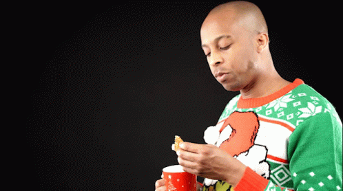 A Man Wearing An Ugly Christmas Sweater Is Holding A Cup And A Piece Of Food GIF