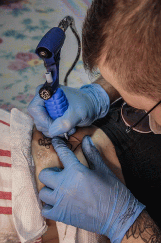 A Man Wearing Blue Gloves Is Getting A Tattoo On Someone 'S Leg GIF