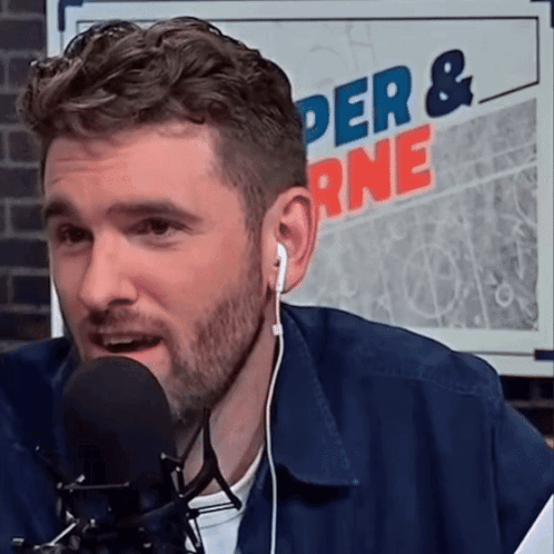 A Man Wearing Ear Buds Is Speaking Into A Microphone In Front Of A Sign That Says Der & Rne GIF