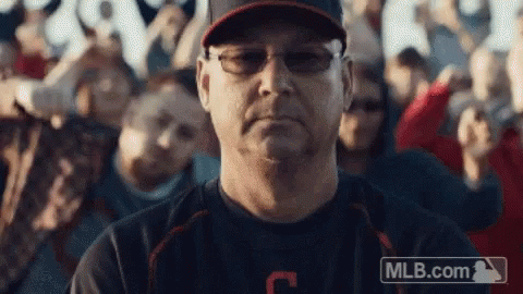 A Man Wearing Glasses And A Baseball Cap Is Sitting In Front Of A Crowd Of People . GIF