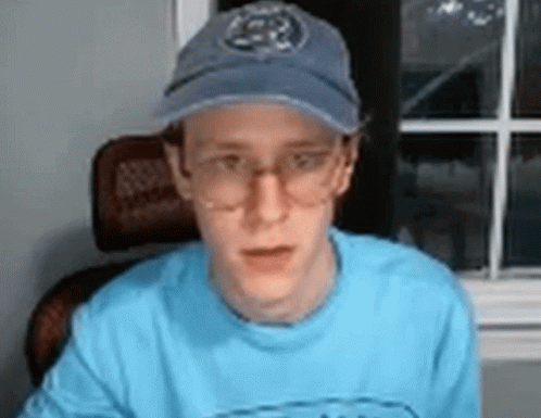 A Man Wearing Glasses And A Baseball Cap Is Sitting In Front Of A Window . GIF