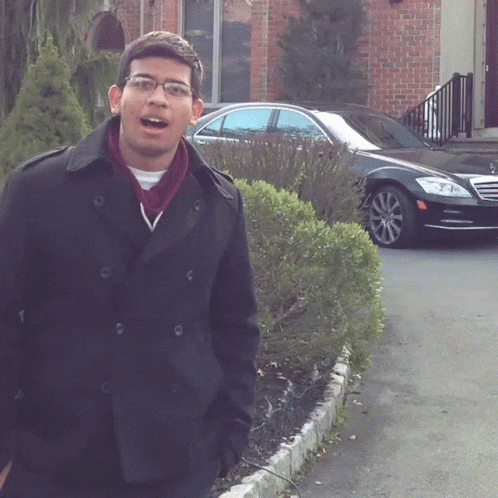 A Man Wearing Glasses And A Black Coat Stands In Front Of A Black Car GIF