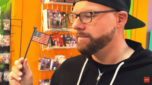 A Man Wearing Glasses And A Black Hat Holds An American Flag GIF