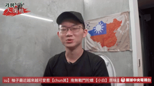 A Man Wearing Glasses And A Black Hat Is Sitting In Front Of A Wall With A Map Of China On It GIF