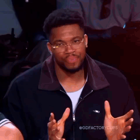 A Man Wearing Glasses And A Black Jacket Is Making A Funny Face While Giving A Thumbs Up . GIF