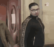 A Man Wearing Glasses And A Black Jacket Is Standing In Front Of A Red Door . GIF