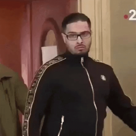 A Man Wearing Glasses And A Black Jacket Is Standing Next To A Door . GIF