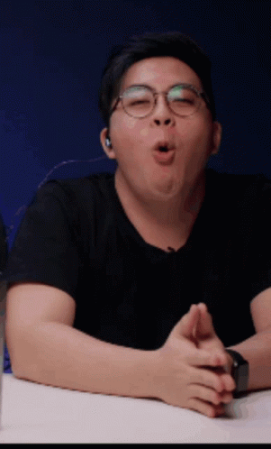 A Man Wearing Glasses And A Black Shirt Is Sitting At A Table With His Mouth Open GIF